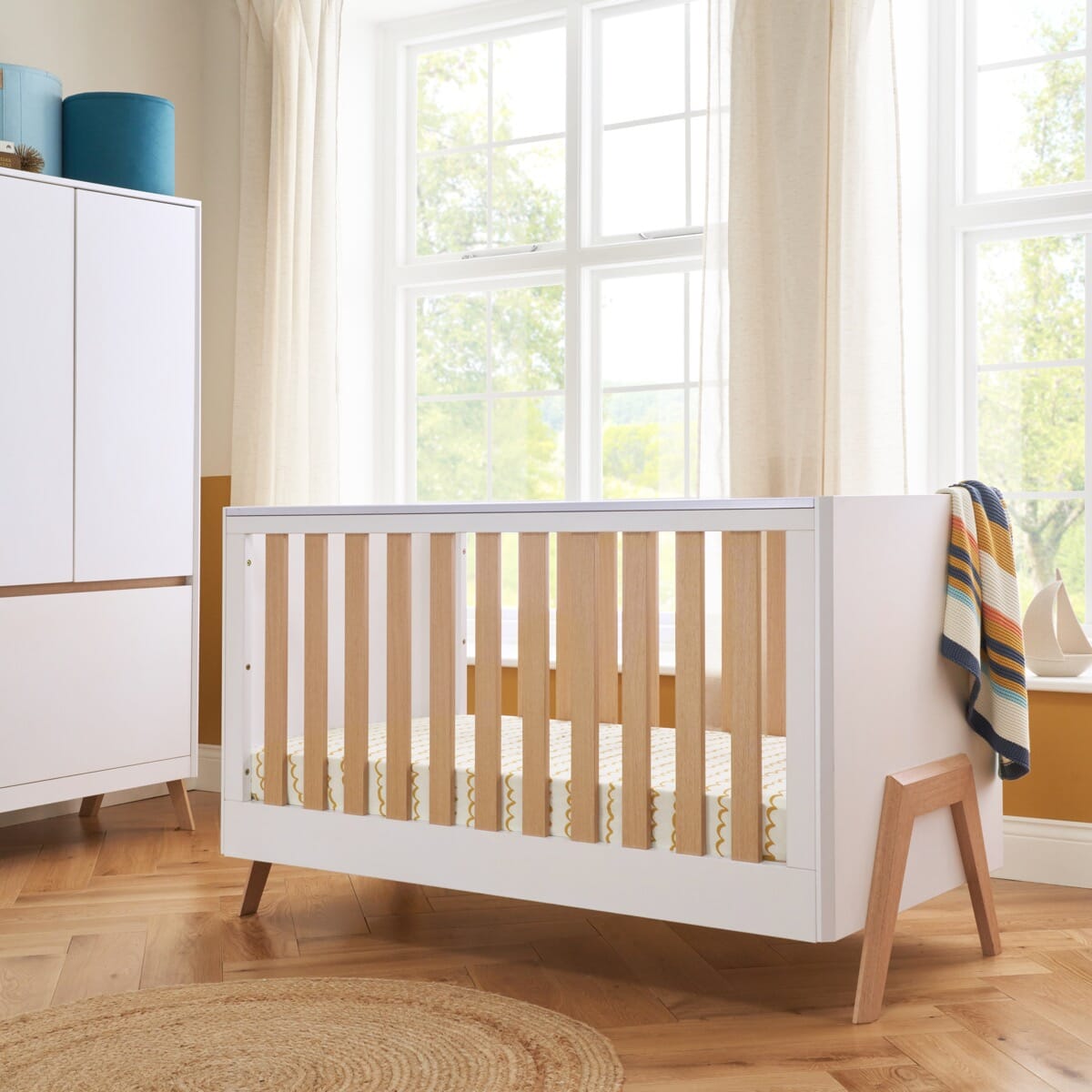 fuori cot bed in white and light oak shown in a brightly lit nursery showing stylish cot design with accompanying wardrobe