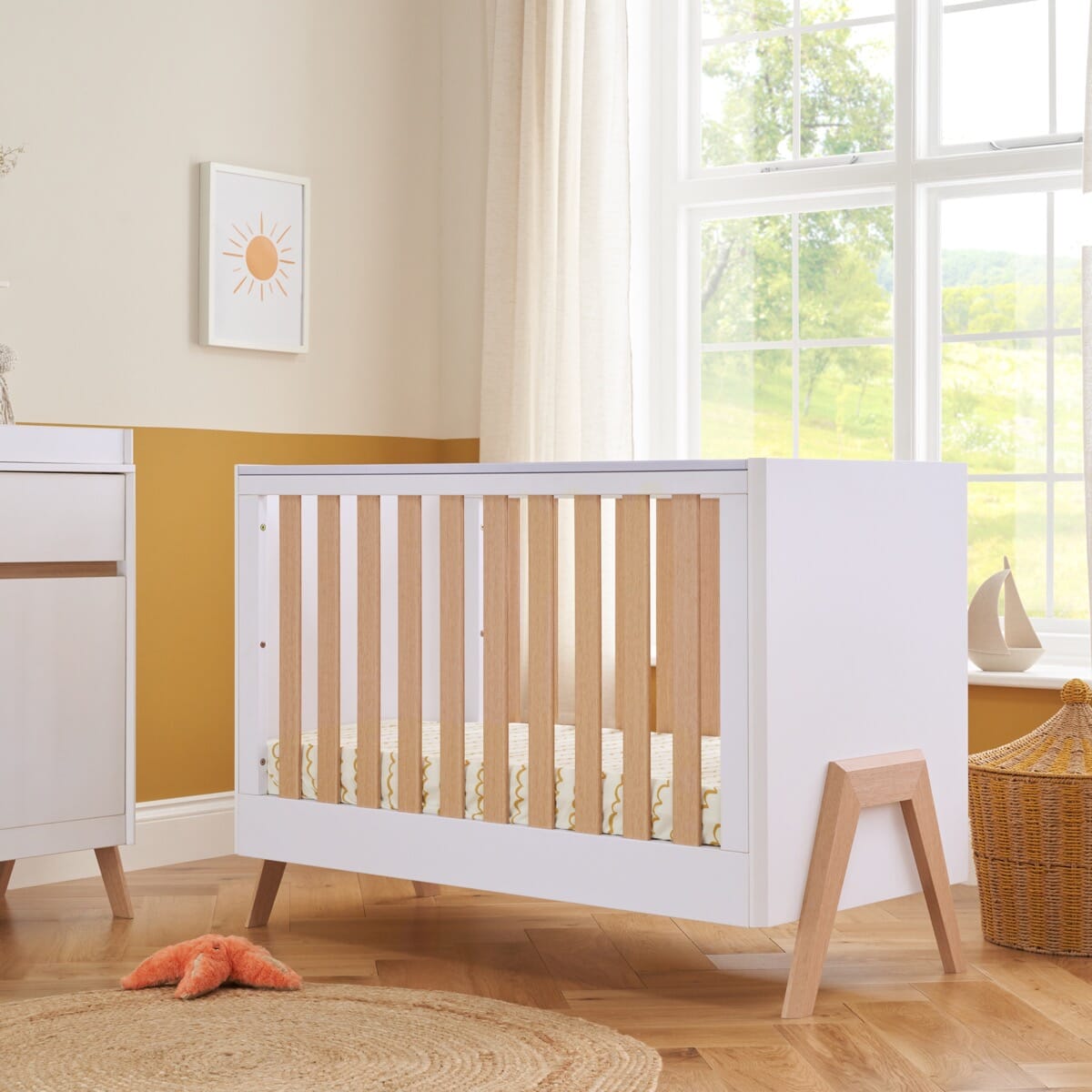 Fuori Mini Cot Bed in white and light oak, placed in a room with a window, next to a wicker basket and a child's toy.