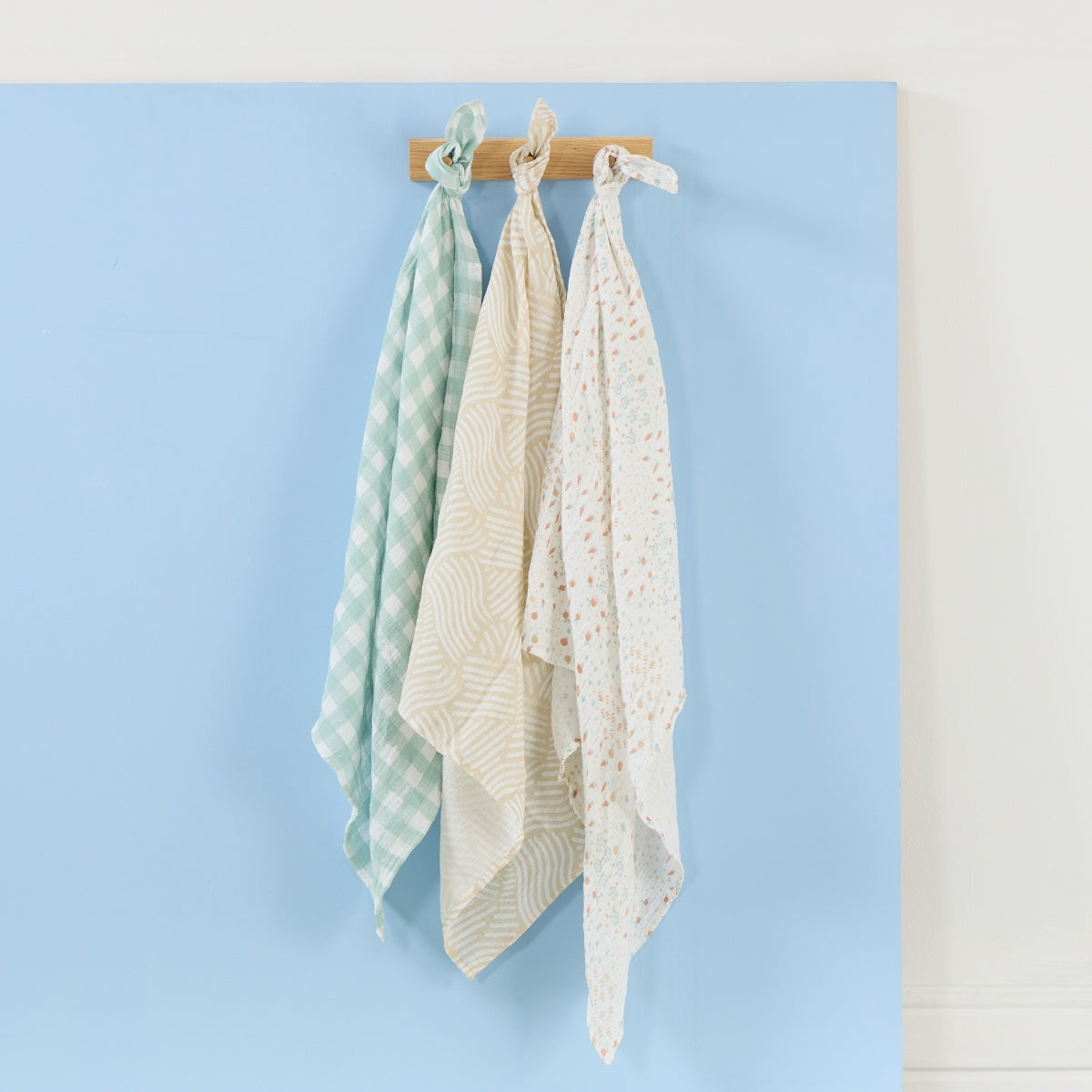 Set of three Good Life large muslin square swaddles hanging on wooden peg rail, featuring soft neutral prints in calming earthy tones