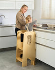 Mum supervises toddler standing safely in the Helping Hands Montessori learning tower in natural finish, perfect for interactive family activities