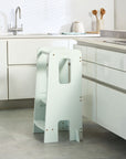 Helping Hands Montessori Toddler Learning Tower in sea mist placed at a kitchen counter, designed to encourage safe toddler participation