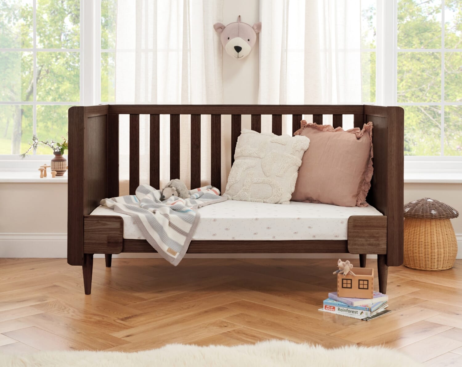 Japandi cot bed in warm walnut shown in sofa bed mode, styled with cushions and soft textiles in a calm nursery setting with natural light
