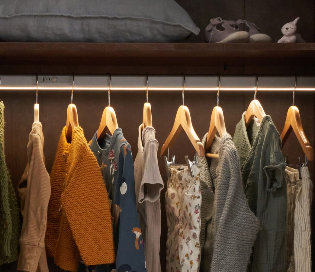 Close-up view inside the Japandi wardrobe in warm walnut, showing the hanging rail illuminated by soft integrated sensory lighting and neatly arranged baby clothes