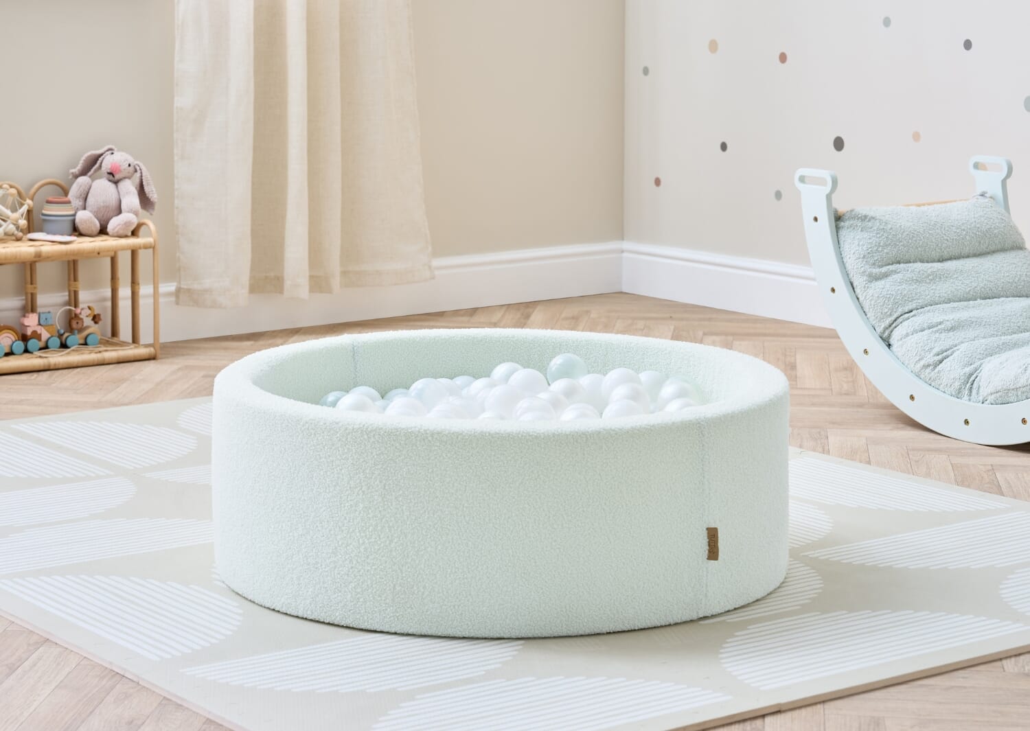 Lifestyle image of the Bola baby ball pit in sea mist styled in a neutral nursery, featuring a soft boucle finish and white balls for calm sensory play