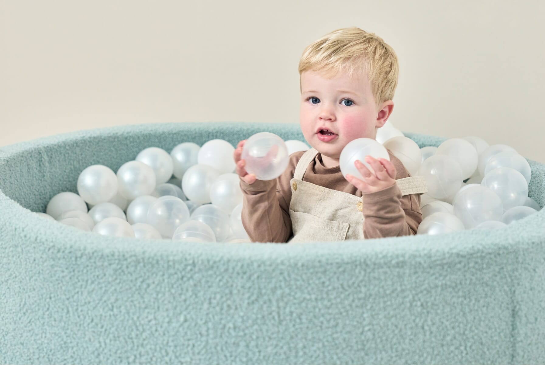 Close-up of a baby holding play balls inside the Bola baby ball pit in ocean stone, highlighting soft fabric sides and sensory play appeal