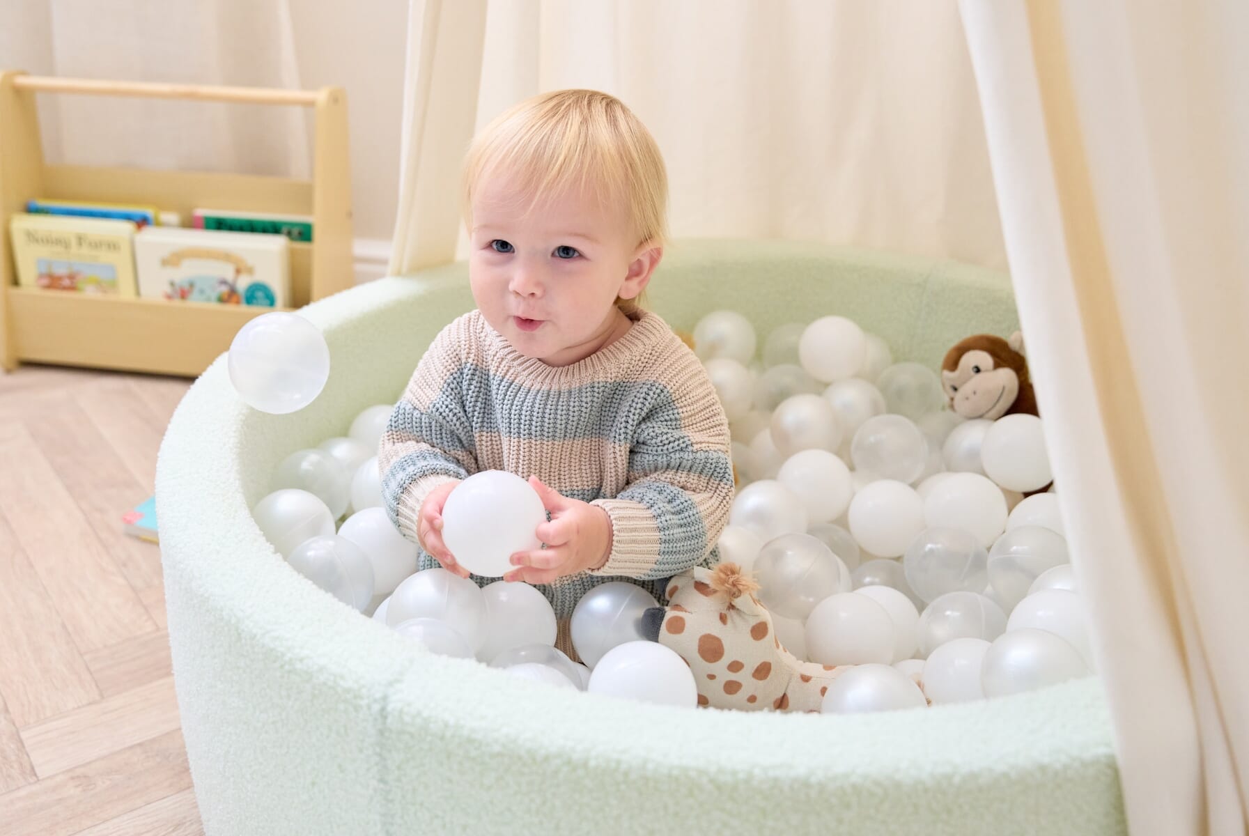 Lifestyle close-up of a smiling toddler enjoying sensory play in the Bola baby ball pit in sea mist, surrounded by soft white balls