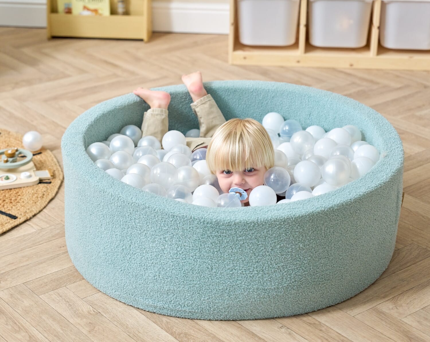Baby relaxing inside the Bola baby ball pit in ocean stone, filled with soft white balls and designed for safe, sensory play