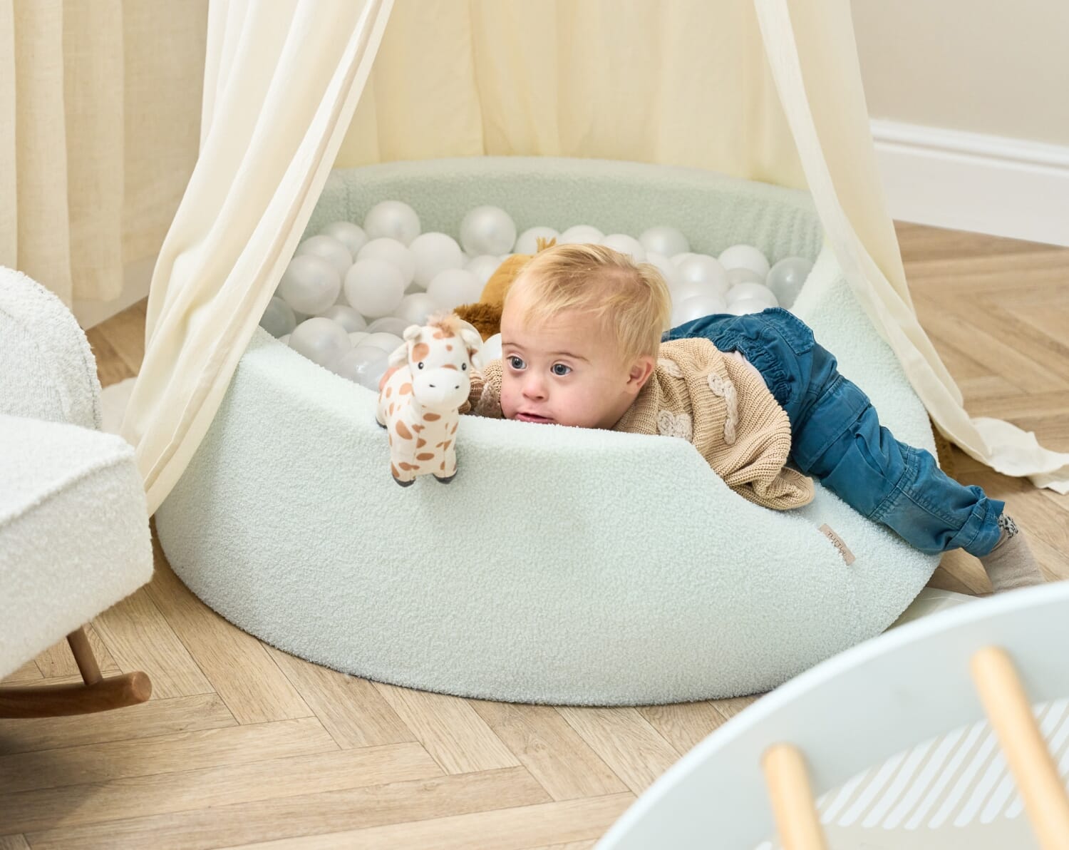 Toddler sitting inside the Bola baby ball pit in sea mist, holding white balls and enjoying gentle sensory play beneath a soft canopy