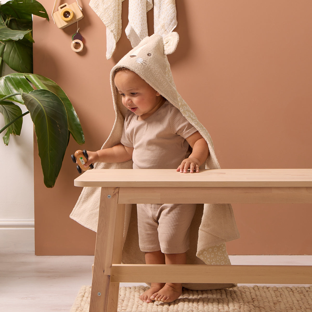 Toddler standing by a wooden bench wearing the Luca Leopard hooded towel, beige terry fabric with playful leopard print inner lining