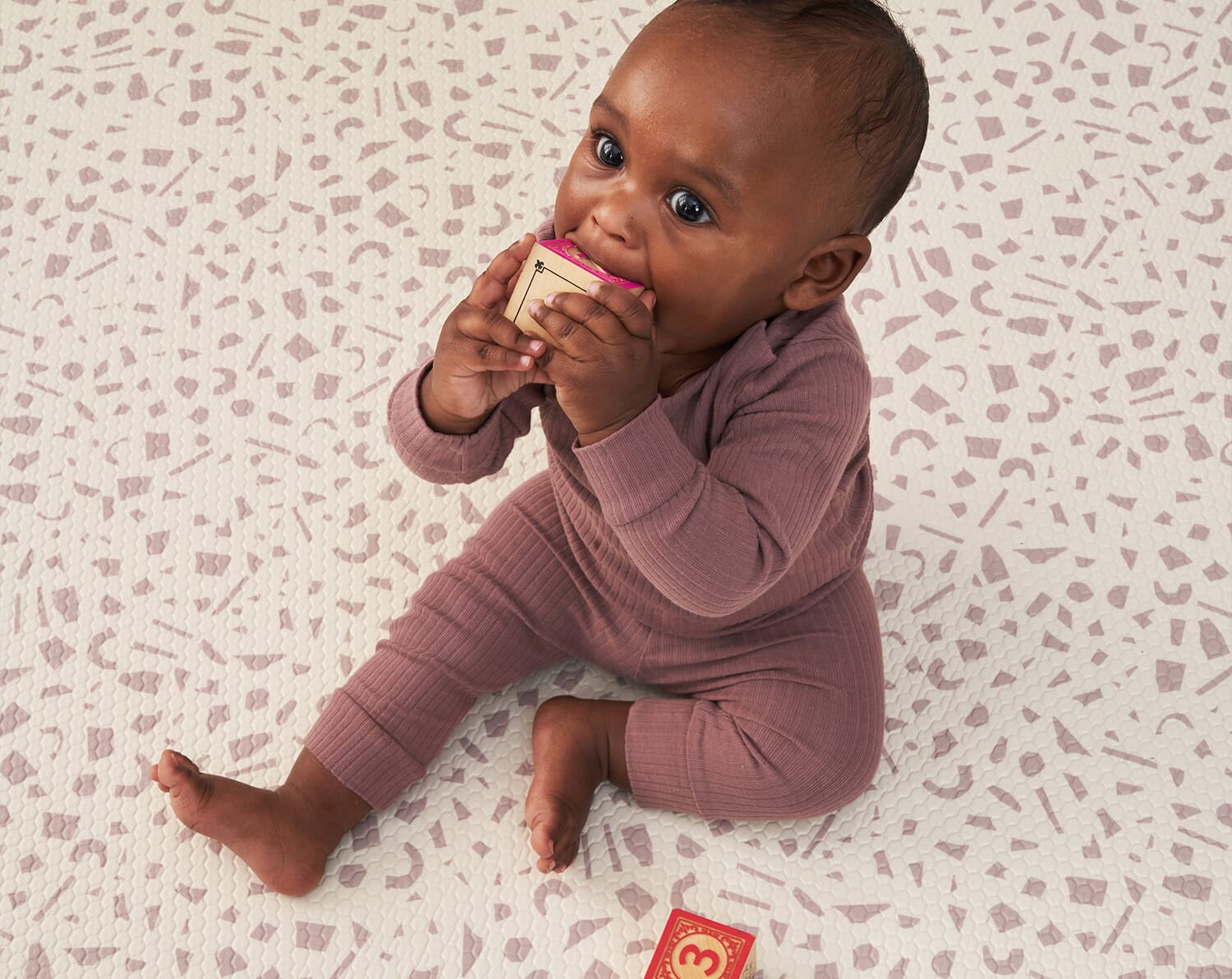 Baby sitting on the Bubble Terrazzo play mat, holding a wooden block to their mouth, showing its cushioned, supportive surface for safe play