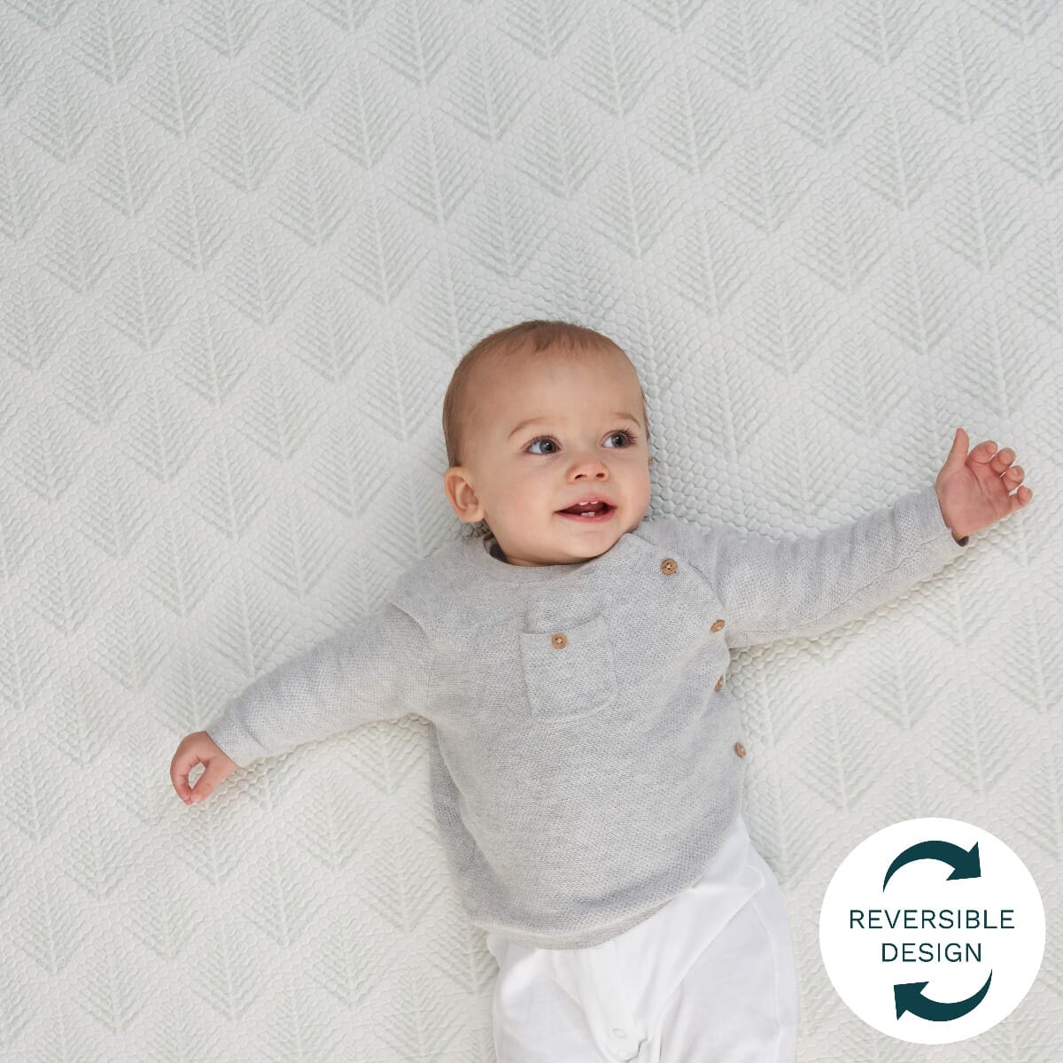 Toddler lying on the white fan-pattern side of the reversible play mat, highlighting its soft padded texture for comfy floor play
