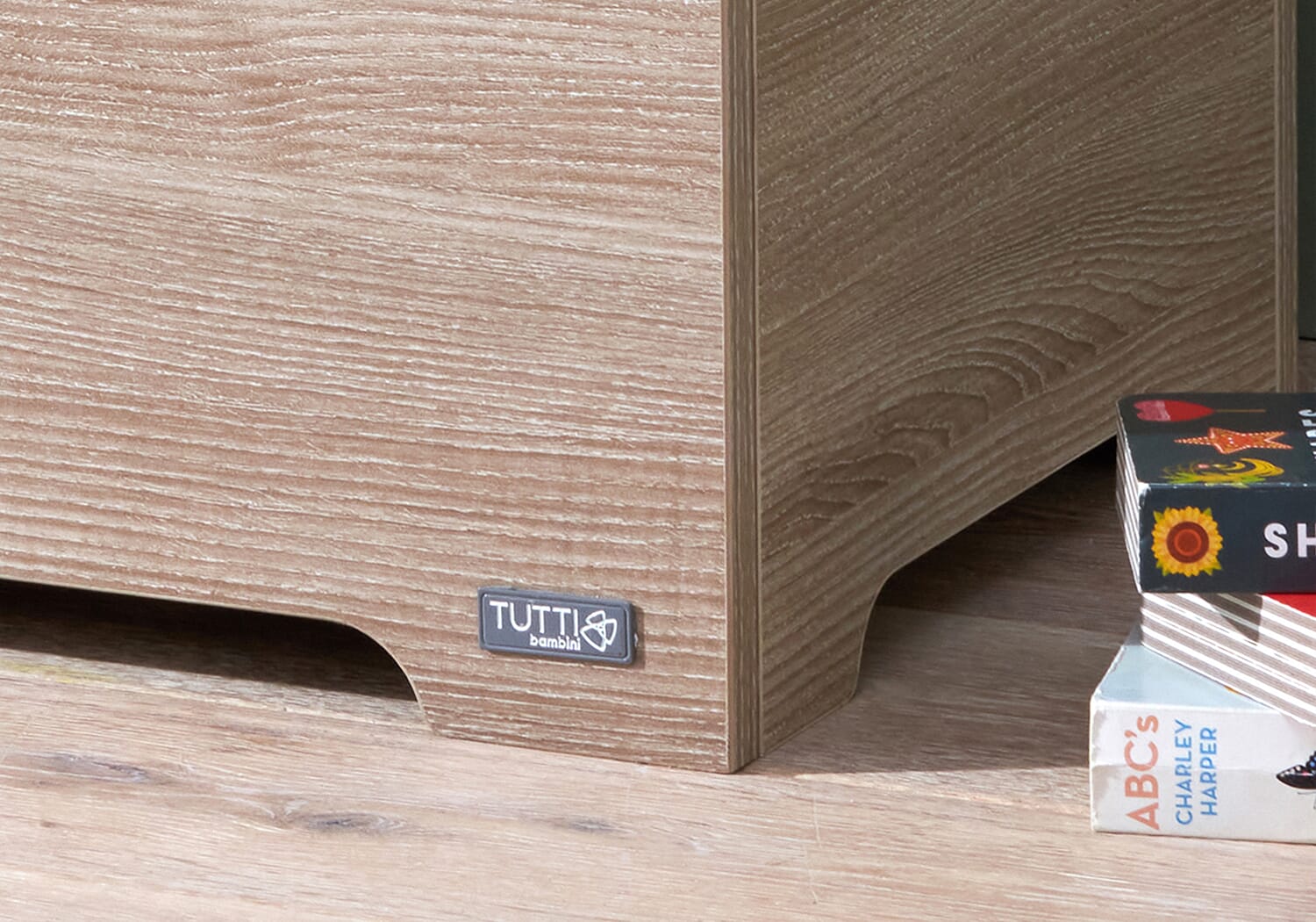 Close-up of the Modena toy box in oak showing the Tutti Bambini logo and textured wood grain detail on the lower corner panel