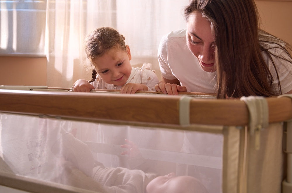 Mum and daughter bonding over newborn in CoZee bedside crib in Scandinavian walnut and ecru, with mesh side panel and soft lighting