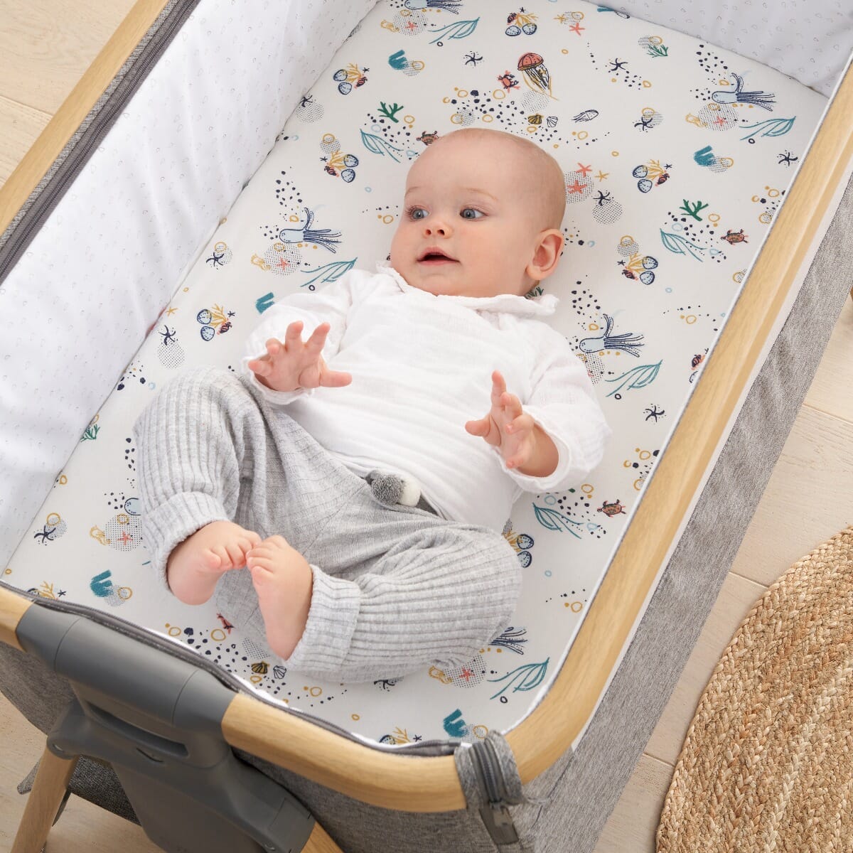 Baby lying in bedside crib on the Our Planet fitted sheet with playful ocean-inspired print, crafted from soft breathable cotton