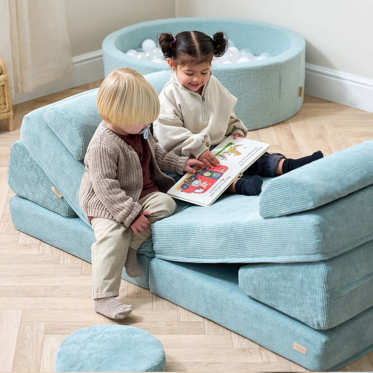 Two toddlers sitting on the Playrama Montessori Kids Play Sofa in ocean stone, sharing a picture book and enjoying quiet reading time