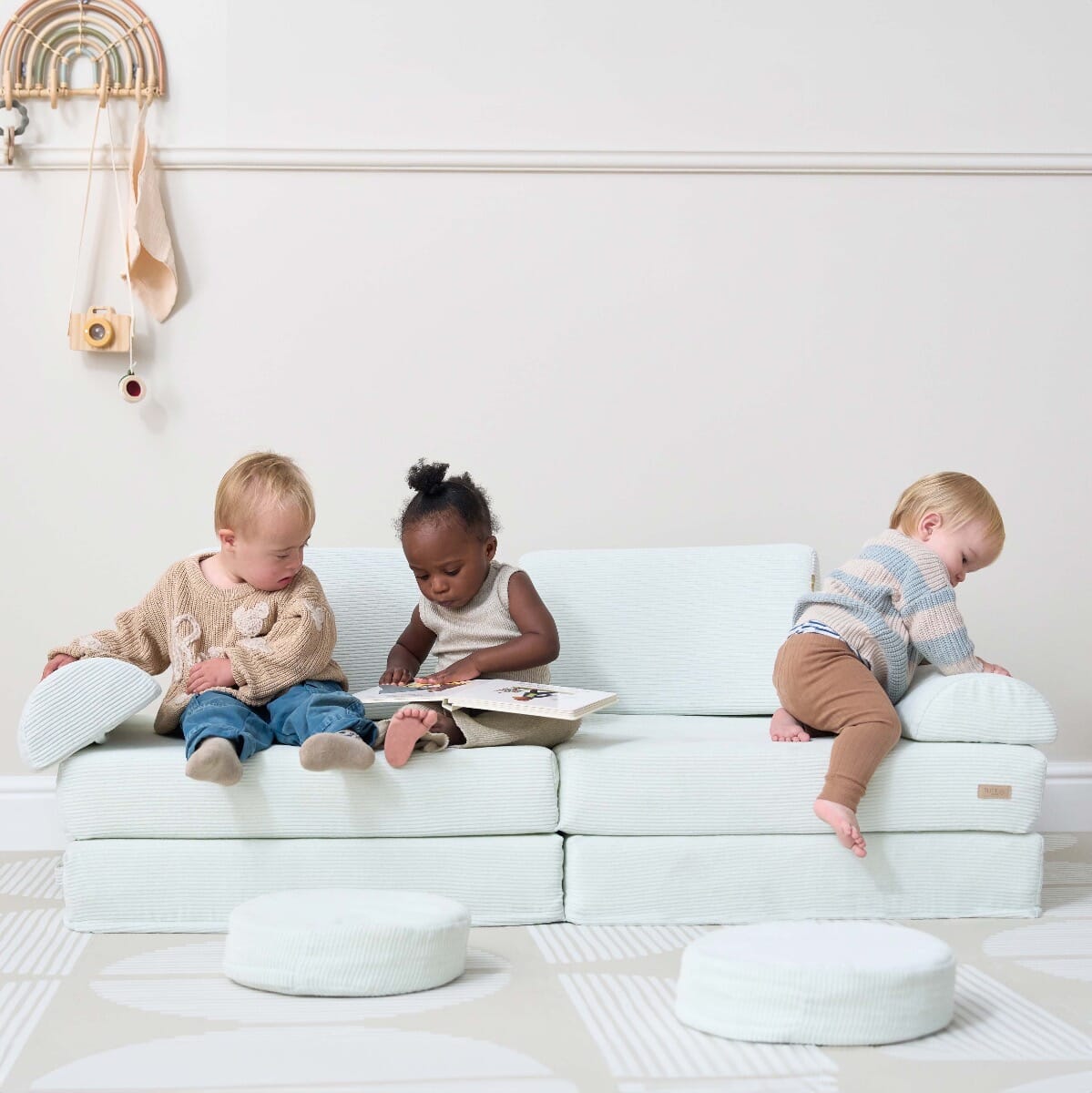 Three toddlers sit and play on the Playrama Montessori kids play sofa in sea mist, with one reading a book and others exploring cushions
