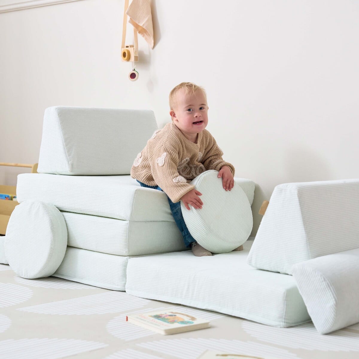 Toddler sits on the Playrama Montessori kids play sofa in sea mist, holding a round cushion while building a playful setup