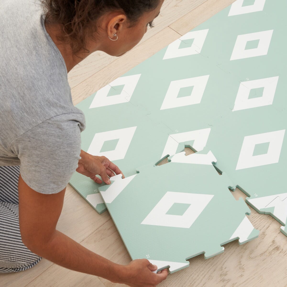 Parent assembling Diamond Mint puzzle playmat tiles on a wooden floor, aligning interlocking pieces to create a soft, cushioned play area