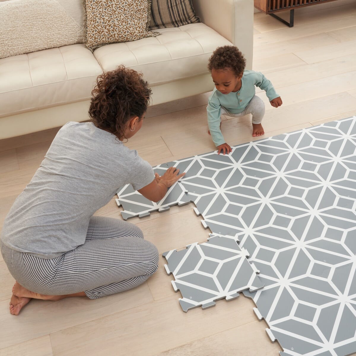 Parent and toddler assembling grey puzzle playmat tiles with geometric pattern, showing easy interlocking design for a soft play space