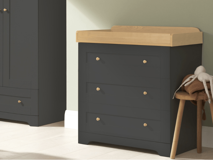 Angled lifestyle view of the Rio chest changer in slate grey with oak top, styled in a calm nursery setting with soft natural light