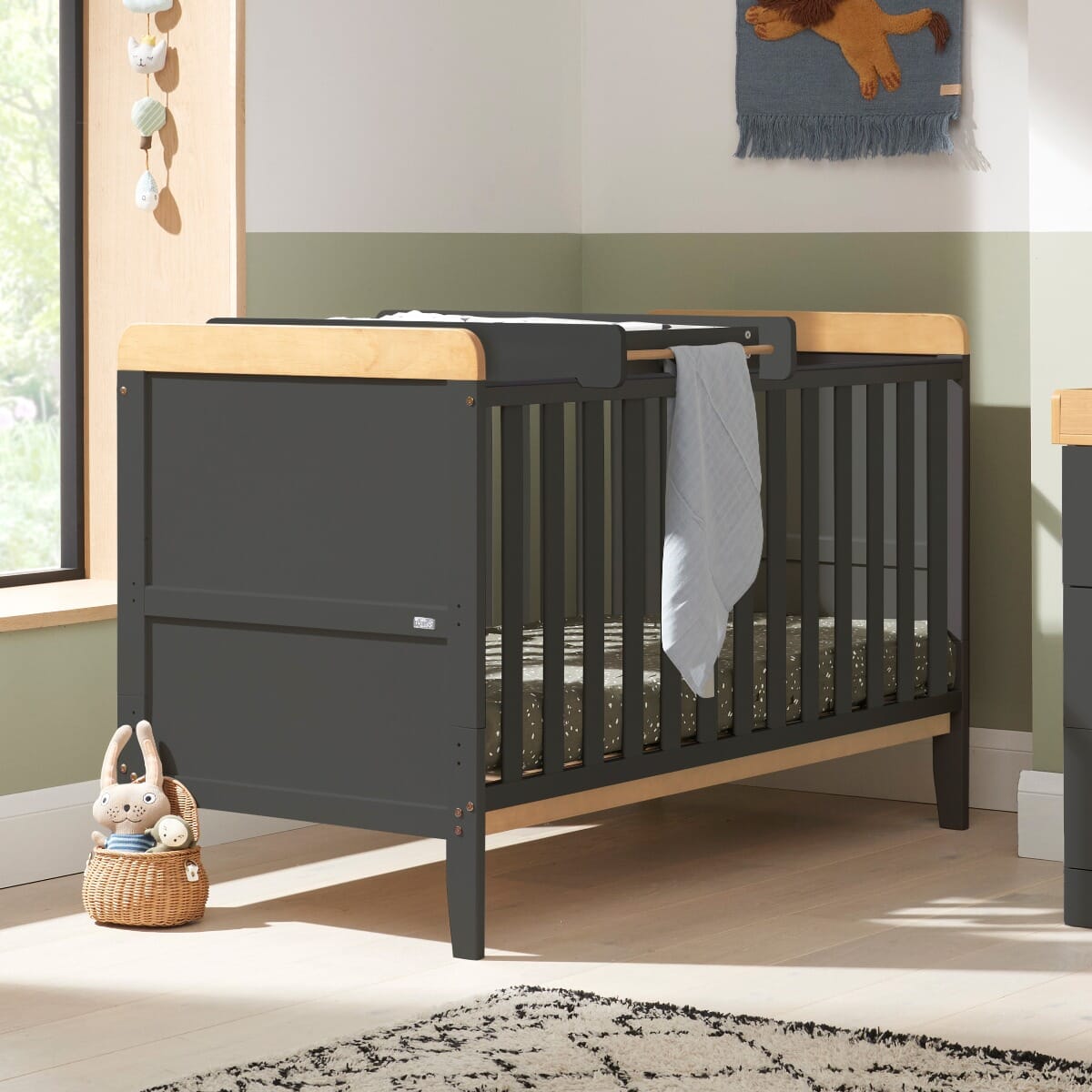 Nursery room view of the Rio cot bed in slate grey and oak, featuring warm wood accents, slatted sides and a calm, modern setting