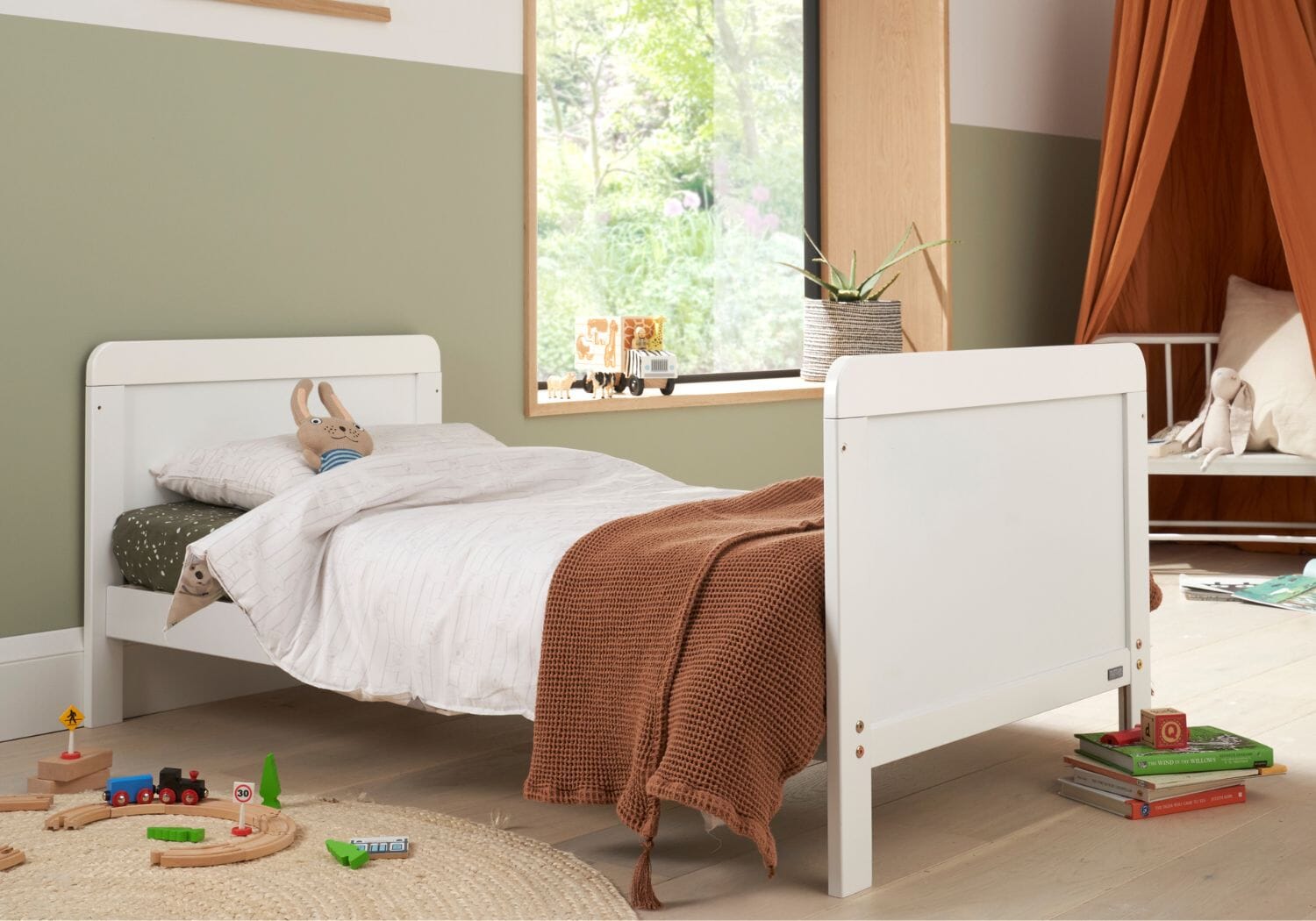 rio cot bed in white shown in toddler bed mode with toys on the floor and large window