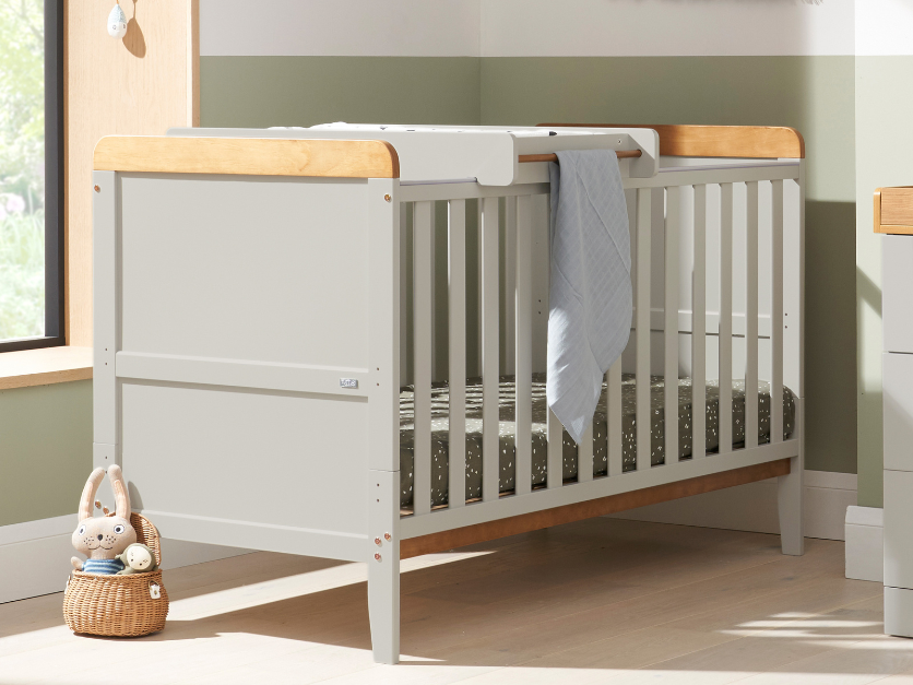 close up of rio cot bed with cot top changer in honey oak and dove grey, featuring hanging towel and rabbit toy in basket