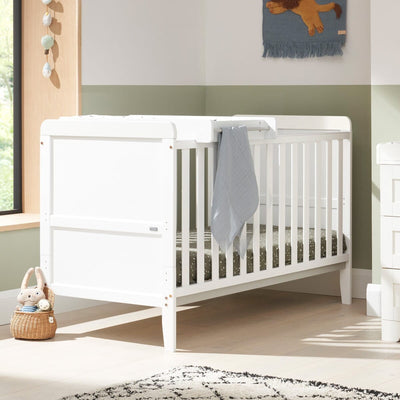 Lifestyle nursery view of the Rio cot bed and top changer in white styled with bedding and accessories for a modern baby room.