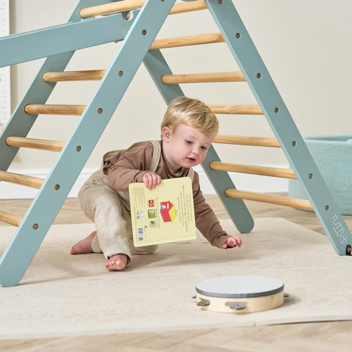 Toddler sits beneath the Scala Montessori climbing frame in ocean stone, holding a book, showing how the frame supports open-ended play and exploration