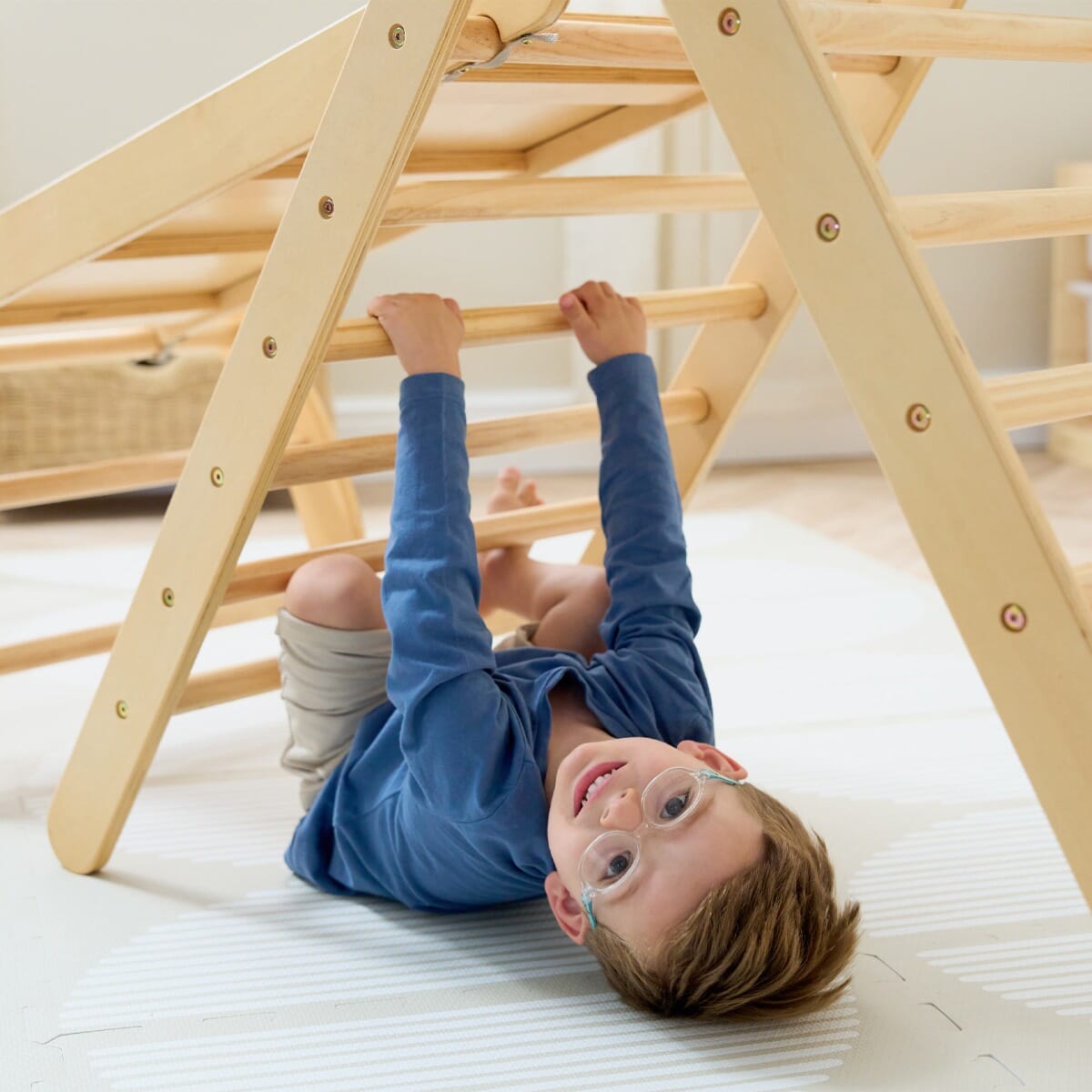 Child smiling while hanging upside down from the Scala Pikler climbing frame and slide in natural wood, promoting active Montessori play