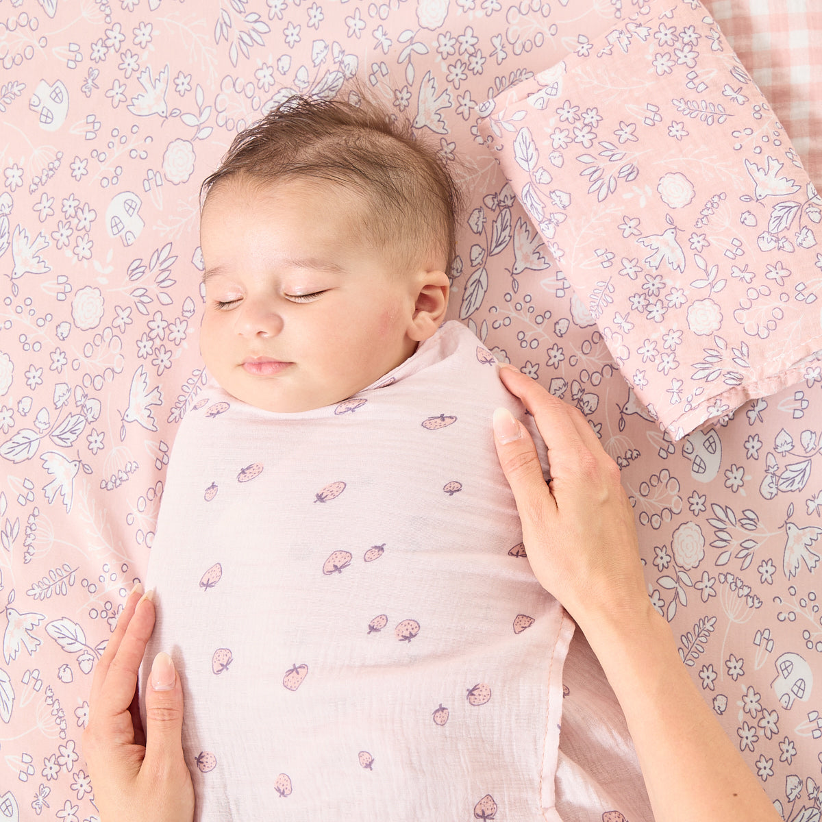 Close-up of baby swaddled in Tiny Tails muslin with berry print, resting on coordinating floral fabric with gentle hand on chest for comfort