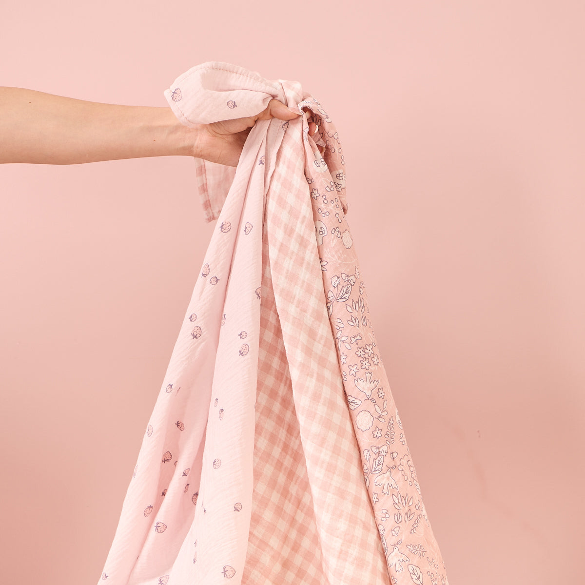 Close-up of three Tiny Tails large muslin square swaddles in coordinating pink patterns, showing soft texture and lightweight breathable fabric