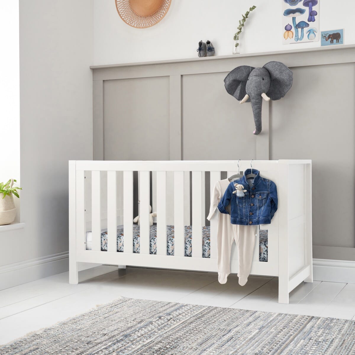 Tivoli cot bed in white styled in a softly lit nursery, featuring slatted sides, neutral decor and everyday baby clothing draped over the rail
