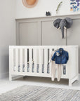 Tivoli cot bed in white styled in a softly lit nursery, featuring slatted sides, neutral decor and everyday baby clothing draped over the rail