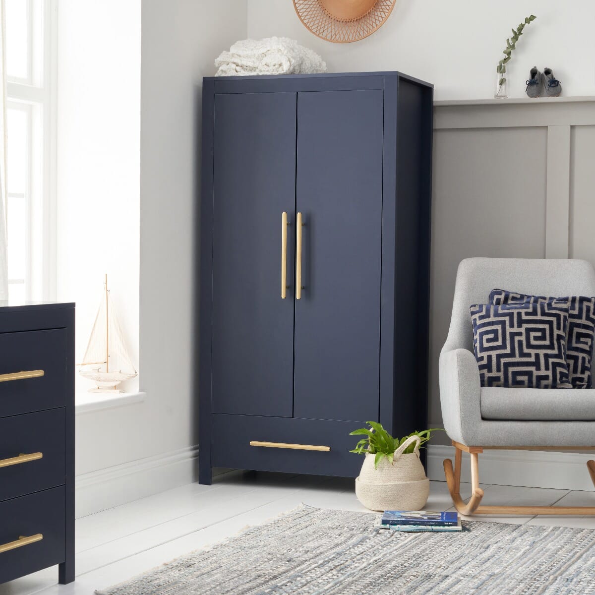 Navy Tivoli wardrobe in a calm nursery setting with gold handles and lower drawer, styled beside a chair and soft accessories