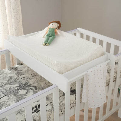 A white Universal Cot Top Changer placed on top of a cot with white bed rails and a patterned bedsheet. A stuffed toy is seated on the mattress.
