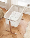 Top view of ZiZee Essential Crib in Oak and Ecru colours, featuring a mesh surrounding, a white mattress, and wooden legs, placed next to a fluffy white rug.